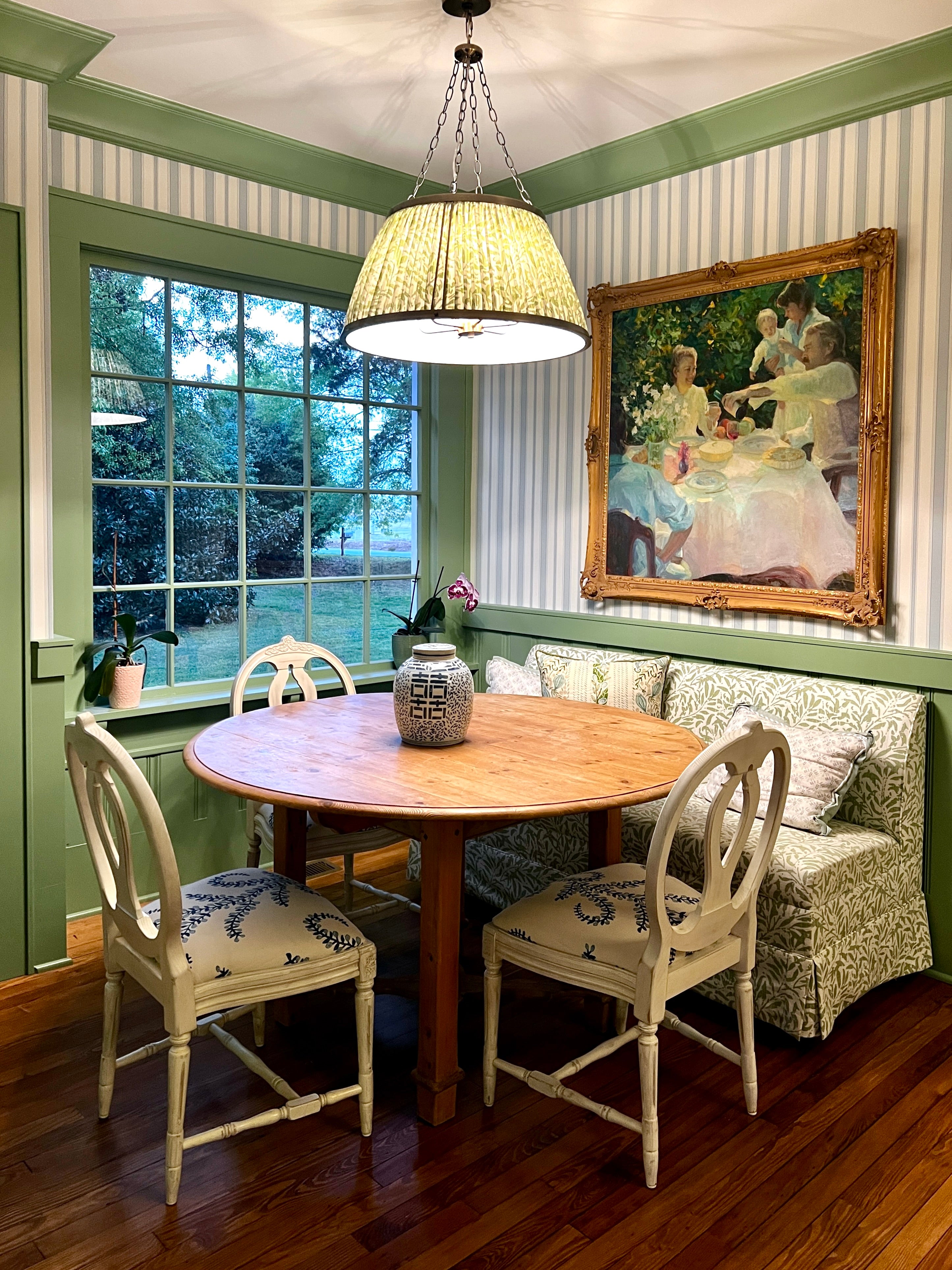 Dining room with wooden table and chairs, large window by Jenny Kramer Decor and decorative painting on the wall. Lee Jofa Stripe wallpaper, Ballard Designs banquette seat and impressionistic oil painting. 